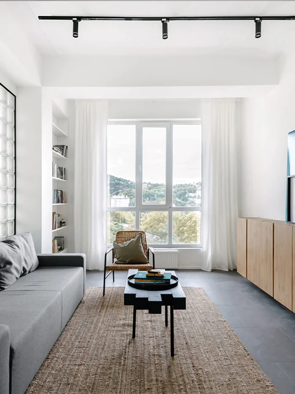 Modern living room with a gray sofa, black coffee table, woven chair, large window, and clean minimalist decor.