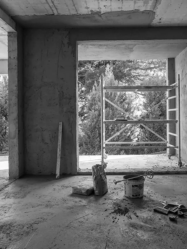 Unfinished interior under renovation with plastering materials, scaffolding by a large window opening, and exposed concrete walls and ceiling.